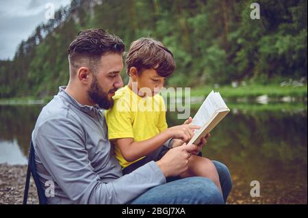 Sur une randonnée, sur la rive de la rivière, le père et le fils lisent un livre Banque D'Images