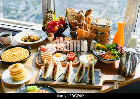 Table avec petit déjeuner continental de rêve - fruits, œufs, sandwichs, salade, boulangerie, boissons chaudes et froides devant la fenêtre Banque D'Images