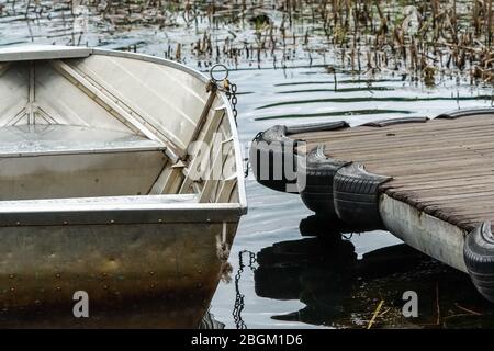 Bateau à rames d'aluminium amarré à une jetée en bois sur un lac avec des ondulations dans l'eau et roseaux dans le concept de fond abstrait plaisir et loisirs d'été Banque D'Images