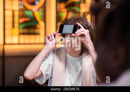 Une fille souriante et curieuse avec son smartphone sur son front assis à la table avec des amis et essayant de deviner un mot écrit sur son écran, gros plan Banque D'Images