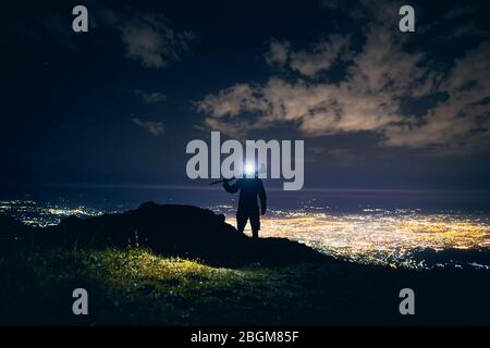 Photographe avec trépied en silhouette marcher sur les montagnes avec lumière LED lumineux contre ciel de nuit avec des étoiles et incandescent ville fond clair. Banque D'Images