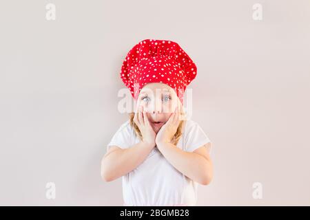 Petite fille blonde souriante dans le chapeau de cuisinier rouge restant sur fond gris neutre et tenant sa joue, surparée, choquée. Espace pour le texte. Banque D'Images