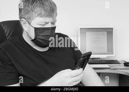 Un adulte dans un masque médical noir regarde soigneusement un smartphone, assis dans une chaise devant un ordinateur. Concept de travail de bureau pendant la quarantaine de Banque D'Images