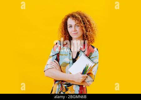 Jolie jeune artiste avec des cheveux bouclés rouges avec bloc-notes et crayons dans les mains Banque D'Images