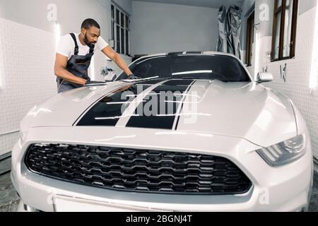 Concept de nettoyage et de détail de voiture. Un professionnel africain du lavage de voiture de qualité, portant des gants et des combinaisons gris, des lingettes et polit la hotte de voiture avec Banque D'Images