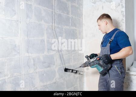 Homme ouvrier du constructeur avec équipement perforateur de marteau perforateur faisant trou dans la construction de mur gris Banque D'Images