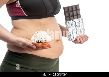 Femme d'âge moyen avec obésité et peau de ventre sagy, avec un cupcake et du chocolat dans ses mains sur un fond blanc, gros plan. Banque D'Images