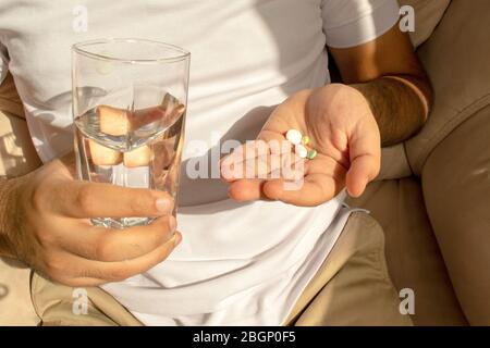 L'homme en t-shirt blanc contient de nombreuses pilules et un verre d'eau dans les mains. Le jeune homme prend des médicaments, des médicaments oraux. Banque D'Images