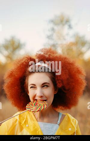 Portrait de femme marche dans le parc dans un imperméable jaune avec des cheveux bouchées d'afro cully Lollipop à la main . Mad View Copy Space concept Halloween Banque D'Images