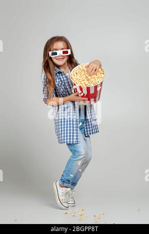 vue sur toute la longueur d'une fille caucasienne souriante avec des lunettes tridimensionnelles rouges et bleues, portant un seau avec du pop-corn sur fond gris de studio. Banque D'Images