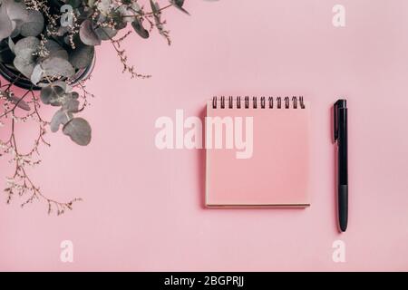 Vue de dessus d'un carnet en spirale rose ouvert, de stylos et d'eucalyptus sur fond de bureau rose. Espace de copie. Banque D'Images