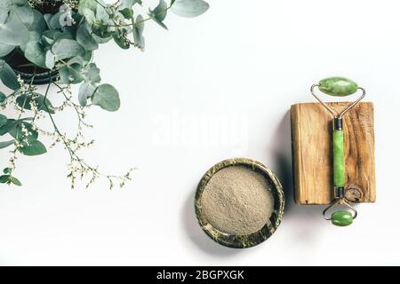 Rouleau de pierre verte sur barre en bois et poudre d'argile dans un bol avec eucalyptus et herbes dans le coin sur fond de papier blanc avec espace de copie. Concept de santé et de beauté. Banque D'Images