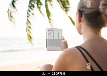 Femme tenant un lecteur électronique à l'extérieur près de la mer avec des palmiers sur fond. Détendez-vous et profitez d'une lecture de livres préférés en plein air dans un voyage. Banque D'Images