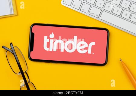 Un smartphone affichant le logo Tinder repose sur un fond jaune avec un clavier, des lunettes, un stylo et un livre (usage éditorial uniquement). Banque D'Images