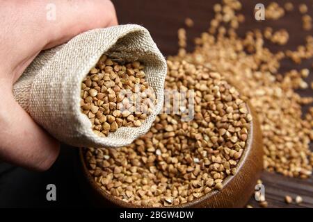 Gruau de sarrasin (graines décortiquées) dans un bol , main avec sac de burlap, fermeture. Ingrédient dans la nourriture pour le petit déjeuner. Sarrasin grains entiers sur table en bois, selecti Banque D'Images