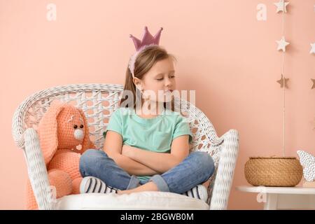 Petite fille indignée avec jouet assis dans un fauteuil près du mur de couleur Banque D'Images