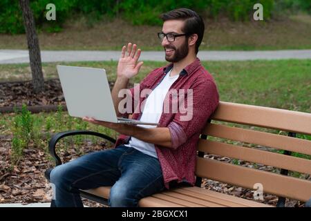 Un jeune homme hipster barbu avec des lunettes se trouve sur un banc extérieur de parc, sur une journée couverte nuageux près d'un sentier de randonnée qui travaille à distance sur son ordinateur portable tryin Banque D'Images