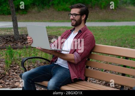 Un jeune homme hipster barbu avec des lunettes se trouve sur un banc extérieur de parc, sur une journée couverte nuageux près d'un sentier de randonnée qui travaille à distance sur son ordinateur portable tryin Banque D'Images