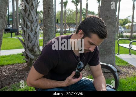 Un homme avec une chemise rouge et un Jean bleu se détend sur un banc de parc jouant avec ses lunettes aviateurs regardant vers le bas sur un beau temps sur un banc de parc Banque D'Images