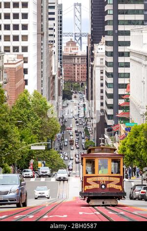 San Francisco, CA - 16 juin 2019 : un téléphérique traditionnel est en cours de fonctionnement au centre-ville sur une rue en pente; au loin, le Bay Bridge Banque D'Images