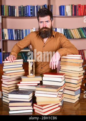 L'homme sur le visage confiant se tient entre des piles de livres, tout en étudiant dans la bibliothèque, les étagères de livres sur fond. Concept de bibliothécaire. Professeur ou étudiant avec des barbes à la table avec des livres, démoutilisés Banque D'Images