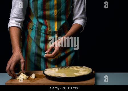 Cuisine de travail. Mains mettant des morceaux de pomme sur le gâteau. Préparation de la cuisson. Faire cuire la tarte aux pommes. Tablier coloré. Ingrédients culinaires. Gâteau aux fruits. Cuisine t Banque D'Images