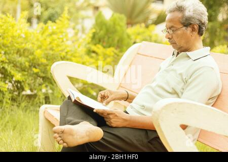 Homme senior au look sain à la fin des années 70 assis dans le jardin à la maison et lire le livre, extérieur - vieil homme se reposant au parc en lisant sérieusement le livre. Banque D'Images