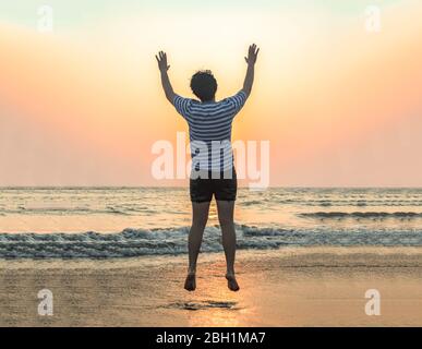 Photo incroyable et parfaitement chronométrée de jeune garçon en bonne santé, sautant dans l'excitation avec les mains dans l'air, après avoir fait sa séance d'exercice de yoga sur la plage. Banque D'Images