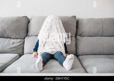 Petite fille assise sur le canapé à la maison se cachant sous une couverture Banque D'Images