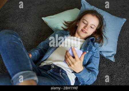 Portrait d'une fille couché sur un tapis à la maison regardant un smartphone Banque D'Images