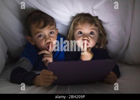 Portrait de frère et de petite sœur allongé côte à côte sous une couverture à l'aide d'une tablette numérique Banque D'Images