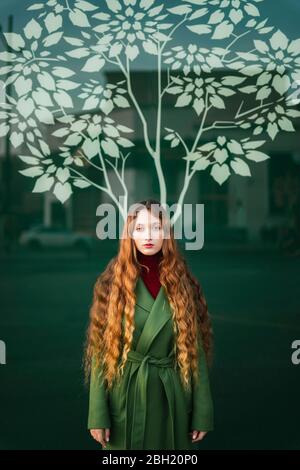 Portrait d'une jeune femme à tête rouge portant un manteau vert debout devant un panneau de verre avec un arbre stylisé Banque D'Images