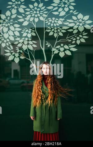 Portrait d'une jeune femme à tête rouge portant un manteau vert debout devant un panneau de verre avec un arbre stylisé Banque D'Images