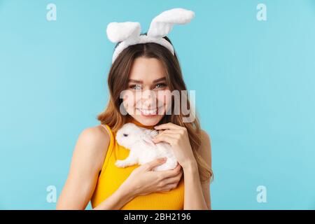 Portrait de la jeune femme heureuse portant des oreilles de lapin jouet souriant et tenant un petit lièvre animé isolé sur fond bleu en studio Banque D'Images