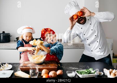 Père joueur avec deux enfants couvrant ses yeux avec des tomates dans la cuisine Banque D'Images