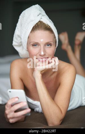 Portrait d'une femme magnifique avec tête enveloppée dans une serviette tenant un smartphone sur le lit Banque D'Images