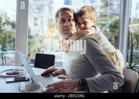 Petit garçon embrassant le père de derrière, tout en travaillant de la maison Banque D'Images