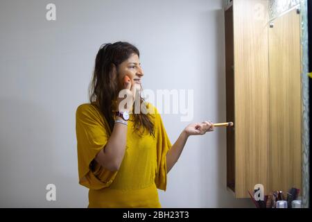 Femme regardant dans le miroir et mélangeant son maquillage tout en tenant un pinceau de maquillage dans son autre main. Banque D'Images