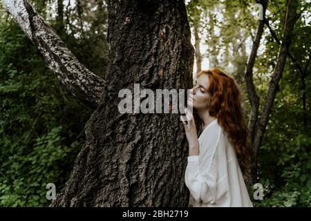 Jeune femme à poil roux embrassant le tronc d'arbre dans la forêt Banque D'Images