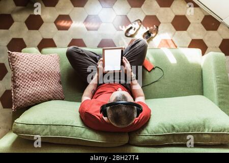 Homme mature assis sur un canapé dans le salon et utilisant une tablette, en écoutant de la musique Banque D'Images