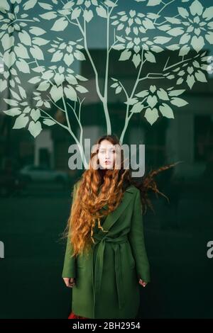 Portrait d'une jeune femme à tête rouge portant un manteau vert debout devant un panneau de verre avec un arbre stylisé Banque D'Images