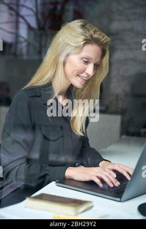 Femme blonde souriante utilisant un ordinateur portable derrière une vitre au bureau Banque D'Images