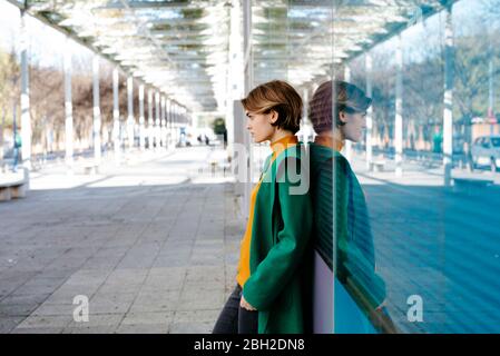 Portrait d'une jeune femme portant un manteau vert et se penchant sur un mur en verre coloré avec son reflet Banque D'Images