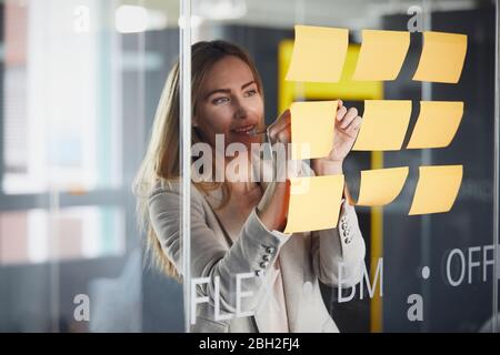 Femme d'affaires écrivant sur des notes adhésives sur un panneau en verre dans le bureau Banque D'Images