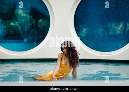 Jeune femme portant une robe jaune d'été assise à la piscine Banque D'Images
