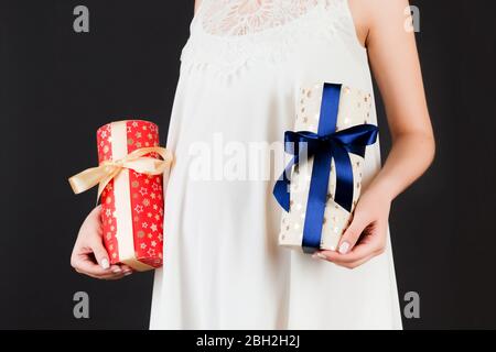 Gros plan sur deux boîtes cadeaux dans les mains de la femme enceinte contre son ventre sur fond noir. Est-ce un garçon ou une fille qui attend des jumeaux. Espace de copie. Banque D'Images