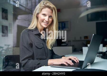 Portrait d'une femme blonde souriante utilisant un ordinateur portable derrière un pare-brise au bureau Banque D'Images