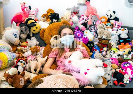 Fille avec masque, assise sur un canapé, couverte de jouets en peluche Banque D'Images