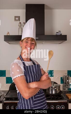 Angleterre Royaume-Uni. 2019. Jolie femme tenant une cuillère en bois et portant un chef blanc chapeau cuisine dans une cuisine moderne. Banque D'Images