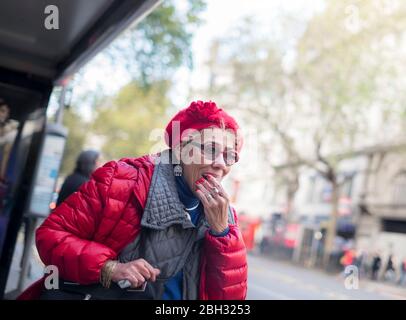 Une femme âgée dans une veste rouge et un chapeau attendant à un arrêt de bus sur EFFRA Road, Brixton, Londres Banque D'Images